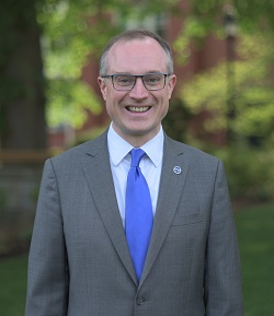 john maura smiling wearing a grey suit with a blue tie.