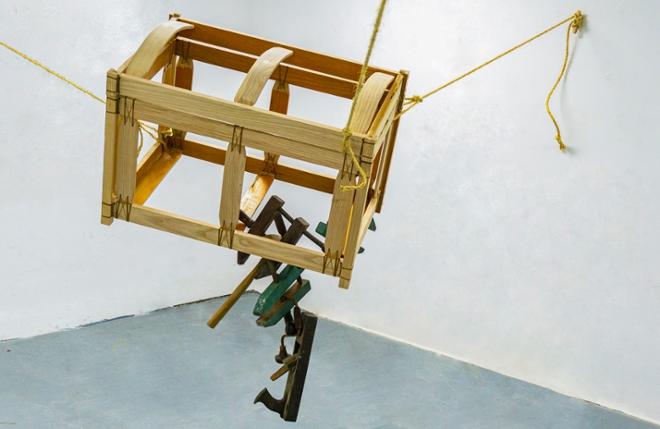 Wooden crate, suspended by three ropes, with woodworking tools hanging down from crate.    