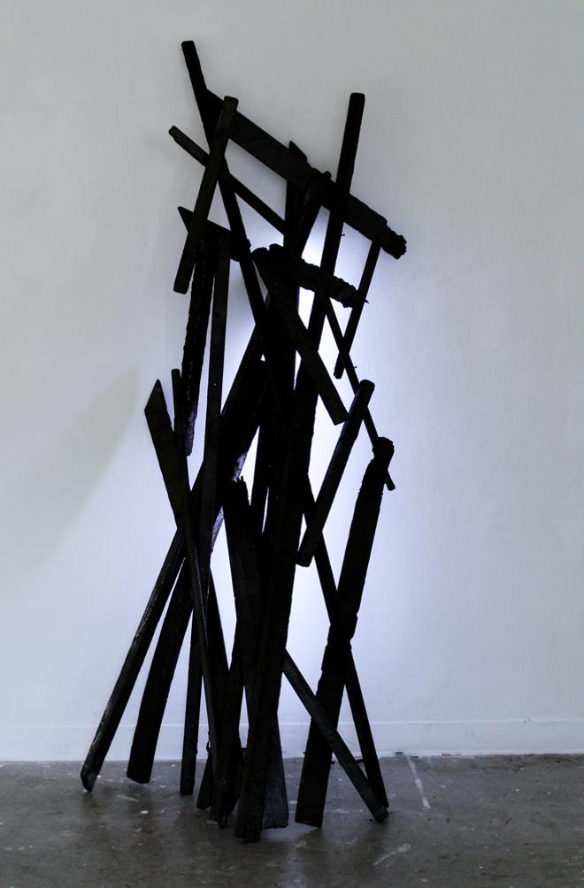 Vertical piled black beams leaning against white wall.