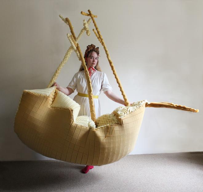 Woman wearing white dress and headband made of bread, holding up boat made of textured foam.