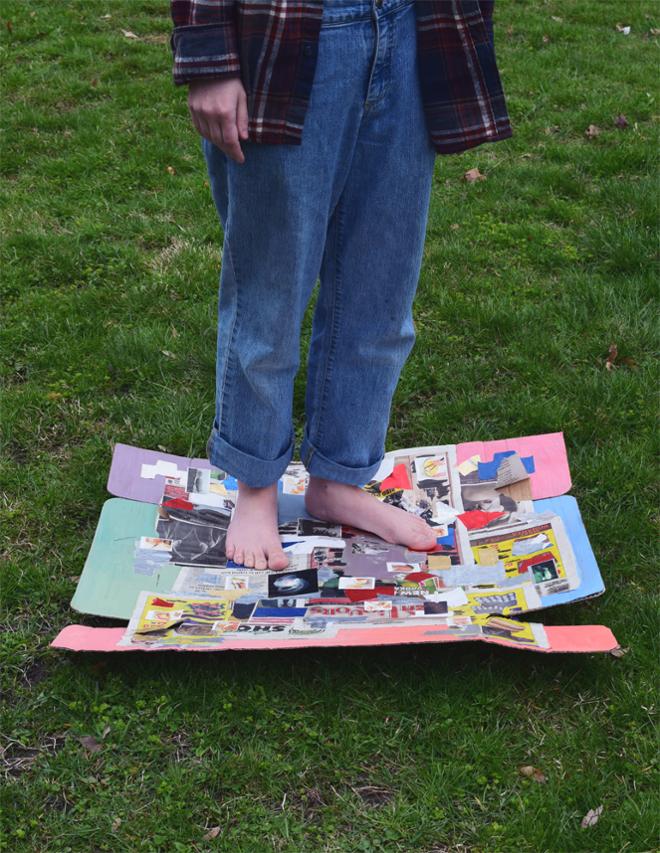 Person wearing jeans standing on cardboard with painted and collaged surface.