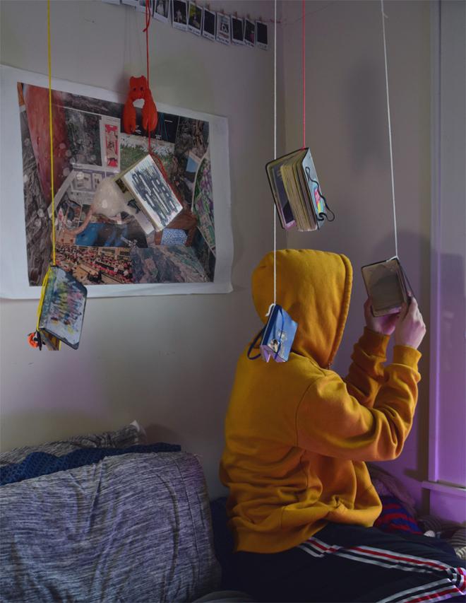 Person in yellow hoodie, sitting on couch, reading a book hanging from a string, surrounded by more hanging books.