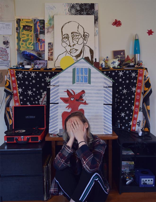 Person sitting on ground with hands covering face, in front of shelves and a makeshift puppet show scene.
