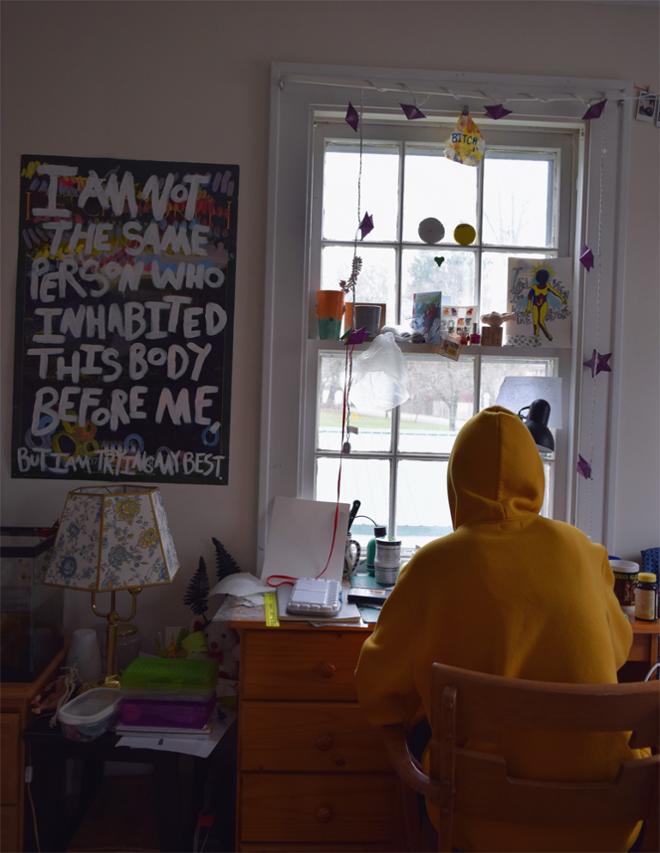 Person in yellow hoodie sitting at cluttered desk in front of window.