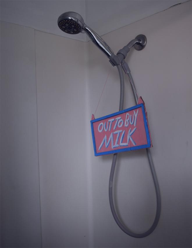 Shower head with pink and blue sign hanging from it saying “out to buy milk”.